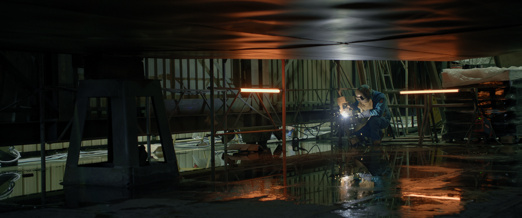 A welder beneath a ship hull with warm reflections