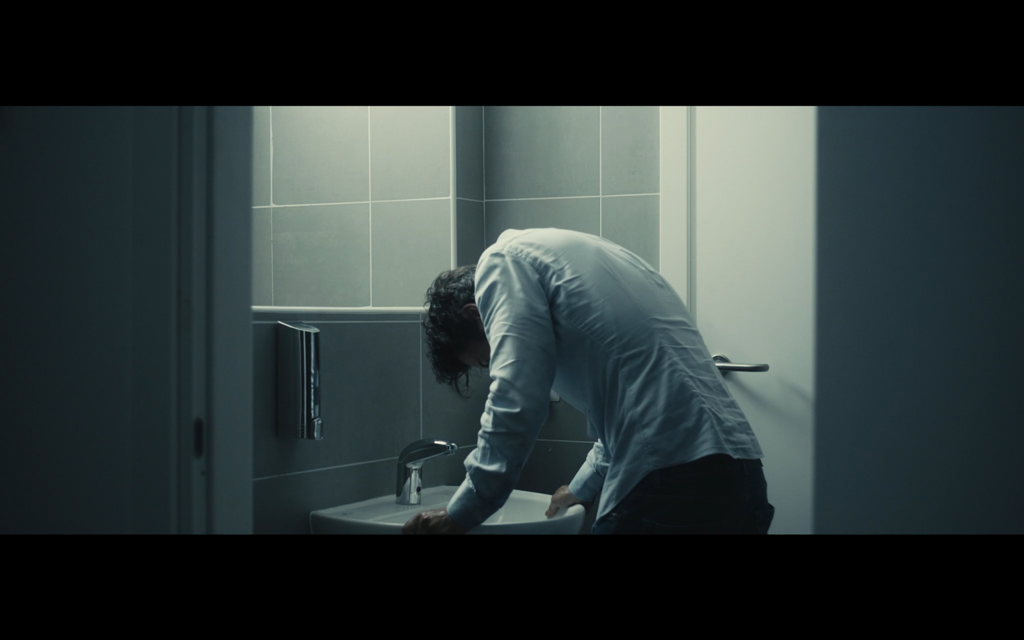 A man leaning over a bathroom sink in cool light